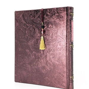 May include: A burgundy journal with an embossed floral pattern. The journal has a decorative spine and a gold tassel bookmark with beads. The cover has a metallic sheen.