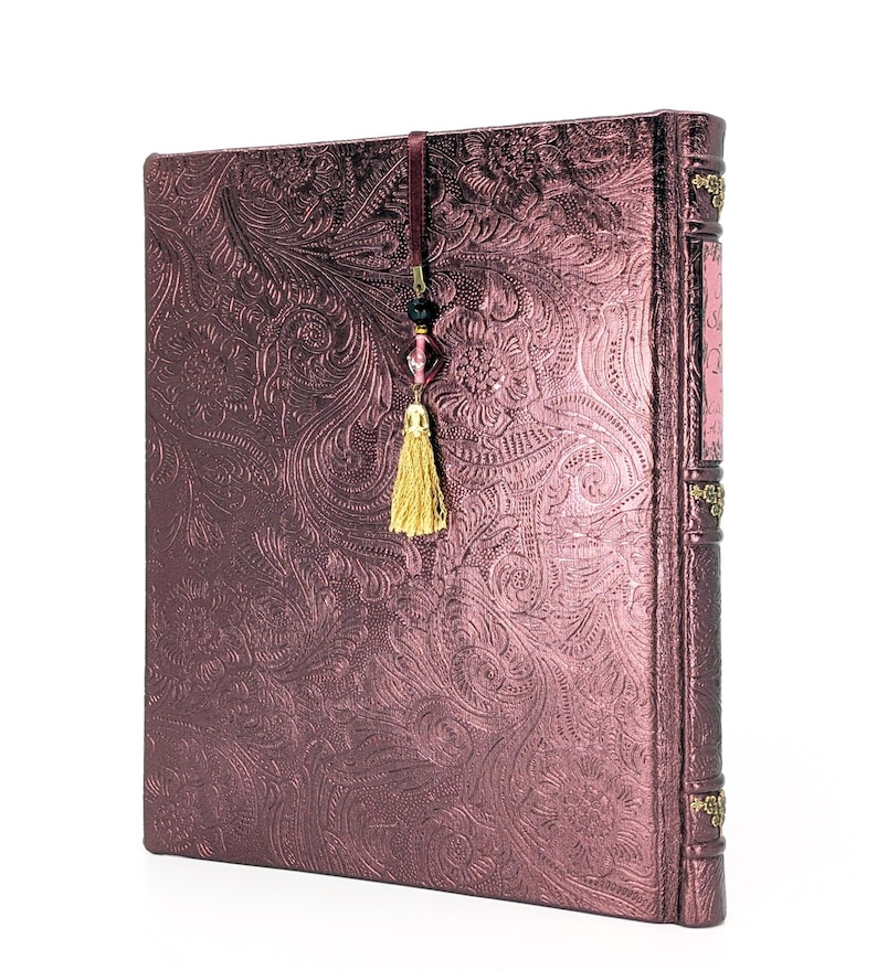 May include: A burgundy journal with an embossed floral pattern. The journal has a decorative spine and a gold tassel bookmark with beads. The cover has a metallic sheen.