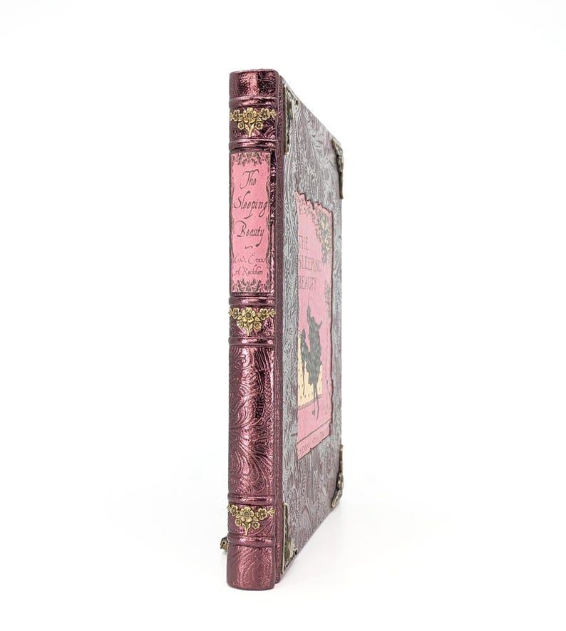 May include: A decorative book titled "The Sleeping Beauty" with a pink cover and spine. The spine has gold accents and the title in elegant script. The cover features a pink illustration and ornate detailing, with metal corner protectors.