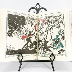 May include: An open book with illustrations displayed on a black metal stand. The illustrations are black and white with accents of red and teal. The book has a burgundy cover with a floral pattern.
