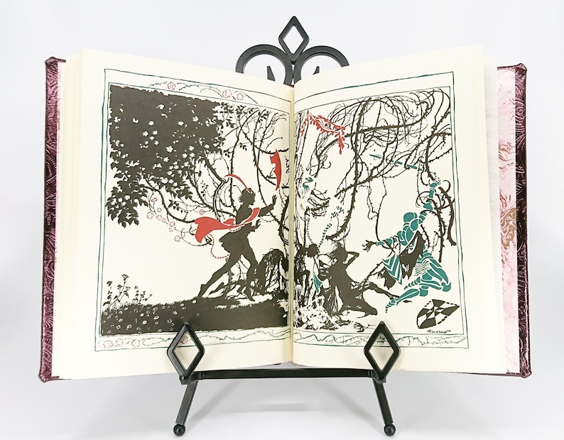 May include: An open book with illustrations displayed on a black metal stand. The illustrations are black and white with accents of red and teal. The book has a burgundy cover with a floral pattern.