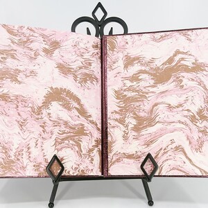 May include: An open book with marbled pages in shades of pink, cream, and brown, displayed on a black metal stand. The book's cover and edges are a deep burgundy color, creating a striking contrast with the patterned interior.