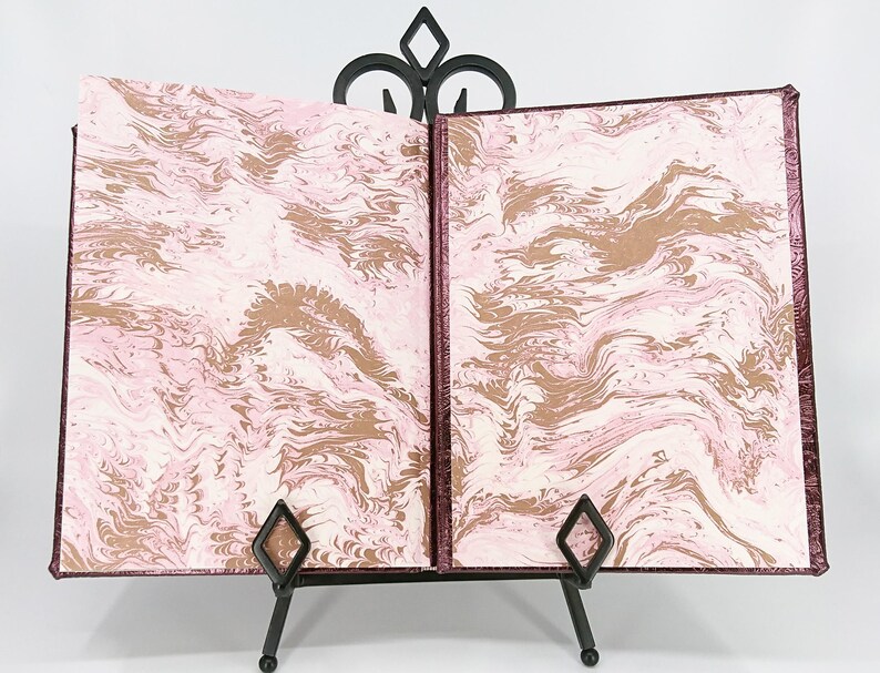 May include: An open book with marbled pages in shades of pink, cream, and brown, displayed on a black metal stand. The book's cover and edges are a deep burgundy color, creating a striking contrast with the patterned interior.