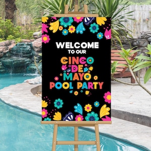 May include: A black sign on a wooden easel with the words "WELCOME TO OUR CINCO DE MAYO POOL PARTY" in colorful text. The sign is decorated with bright floral designs in pink, yellow, blue, and white. The background shows a pool and greenery.