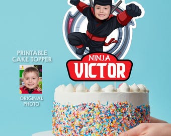 Może przedstawiać: Personalizowana dekoracja na tort przedstawiająca postać ninja z wkomponowaną twarzą dziecka. Dekoracja zawiera czerwony baner z napisem "NINJA VICTOR" i jest umieszczona na okrągłym torcie pokrytym kolorową posypką cukrową.