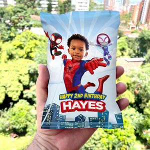 K&ouml;nnte beinhalten: Ein blauer und wei&szlig;er Chip-Beutel mit einem Spiderman-Design. Der Beutel zeigt einen jungen Jungen in einem Spiderman-Kost&uuml;m, einen roten und schwarzen Spiderman und einen wei&szlig;en und violetten Spiderman. Der Beutel tr&auml;gt den Schriftzug "Happy 2nd Birthday Hayes".