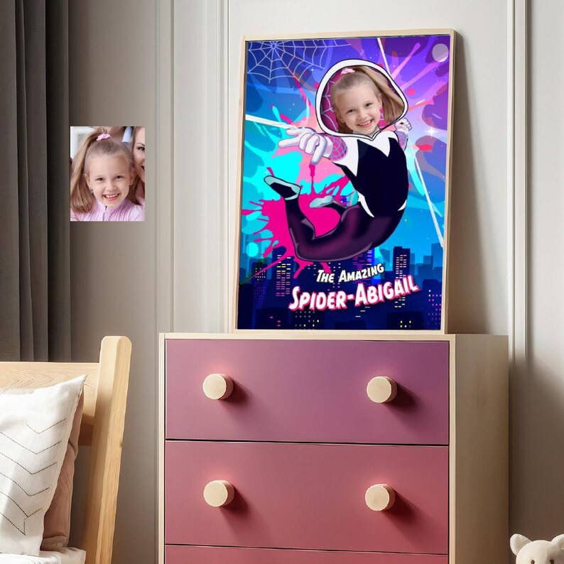 May include: A framed poster featuring a young girl as Spider-Gwen, with the text "THE AMAZING SPIDER-ABIGAIL." The artwork has a vibrant color scheme of pink, blue, and purple. A smaller framed photo of the girl is also visible.