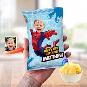 Op de afbeelding: Een gepersonaliseerde chipszak met een kind als Spider-Man, met de tekst "Happy 5th Birthday Matthew". De zak heeft een blauw waterspattenontwerp en bevat een kleine foto van het kind. Een kom met chips staat op de voorgrond.