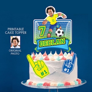 May include: A football-themed cake topper featuring a child in a yellow and blue jersey, a football, a goal net, and the name DECLAN. It includes a large number 7 and two foam finger cutouts labelled #1, all displayed on a white frosted cake.