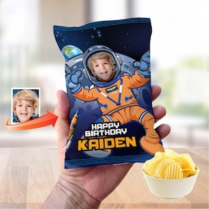 May include: A personalized chip bag featuring a child in an astronaut suit against a space background. The bag reads "HAPPY BIRTHDAY KAIDEN" in white and yellow. A small photo of the child is on the left. A bowl of chips is on the right.