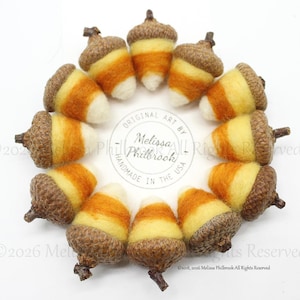 May include: A circular arrangement of twelve felted wool acorns featuring a candy corn color pattern of yellow, orange, and white sections with natural brown acorn caps. The center contains a circular stamp reading Original Art by Melissa Philbrook.