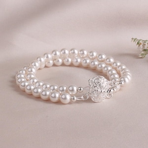 May include: A double-strand pearl bracelet with a decorative silver clasp. The bracelet features numerous round, white pearls. The clasp is intricately designed with a floral pattern. The bracelet is displayed on a soft, neutral-toned surface.
