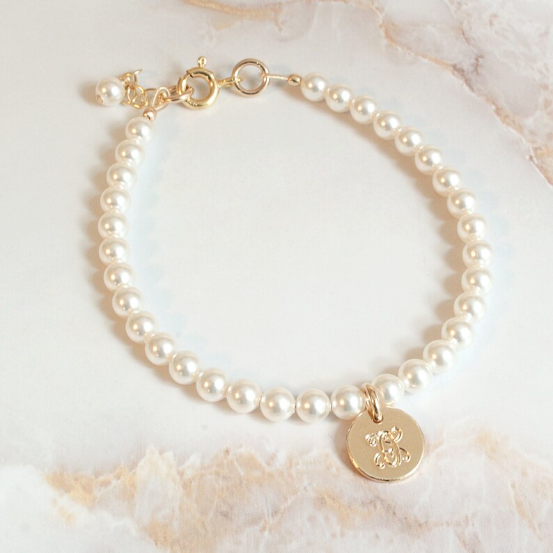 May include: A gold pearl bracelet with a gold charm featuring a monogram. The bracelet is made of small white pearls and has a gold clasp.
