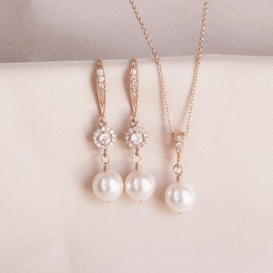 May include: A rose gold jewelry set featuring a necklace and a pair of earrings. The earrings and necklace pendant have a circular design with clear crystals surrounding a pearl. The earrings have a long, slender design with crystals.