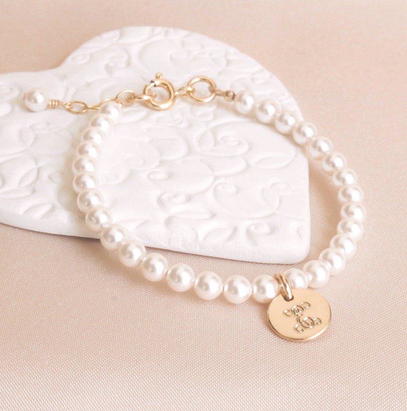 May include: A delicate gold bracelet with a pearl chain and a small gold charm with a monogram. The bracelet is on a white heart-shaped surface.