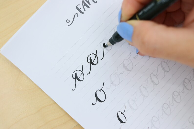 Puede incluir: Una mano escribiendo la letra "o" en cursiva con un bol&iacute;grafo negro sobre papel rayado. El papel tiene l&iacute;neas grises tenues y la palabra "fancy" est&aacute; escrita en la parte superior de la p&aacute;gina.