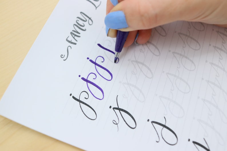 Puede incluir: Una persona practicando caligraf&iacute;a con una pluma morada en un papel rayado. El papel tiene las palabras "Fancy Lettering" escritas en la parte superior. La persona est&aacute; escribiendo la letra "g" en cursiva.