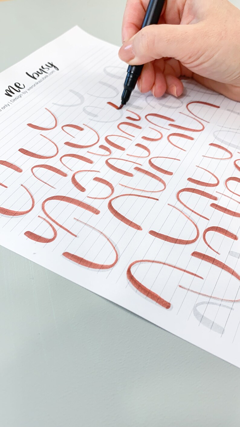 K&ouml;nnte beinhalten: Nahaufnahme einer Hand, die einen schwarzen Marker verwendet, um das Schreiben des Buchstabens "u" in Kursivschrift auf einem Blatt liniertem Papier zu &uuml;ben. Der Buchstabe "u" ist in kupferfarbener Tinte geschrieben.