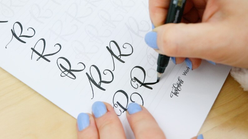 May include: A person practicing calligraphy with a black marker pen on a white sheet of paper. The paper has faint guidelines for the letters. The person is writing the letter "R" in cursive. The text "Twirler Work" is written in cursive at the bottom of the page.
