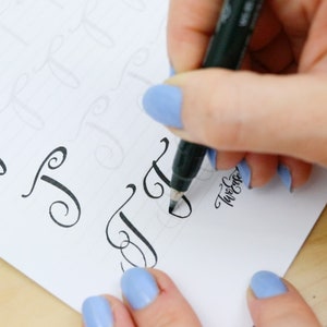 May include: A person's hand is writing a cursive letter 'J' with a black marker on a white sheet of paper. The paper has several other cursive letters written on it. The person's hand is wearing blue nail polish.
