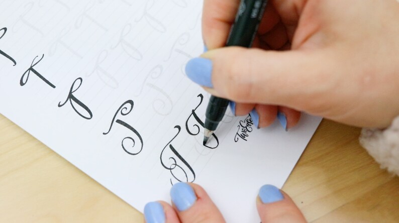 May include: A person's hand is writing a cursive letter 'J' with a black marker on a white sheet of paper. The paper has several other cursive letters written on it. The person's hand is wearing blue nail polish.