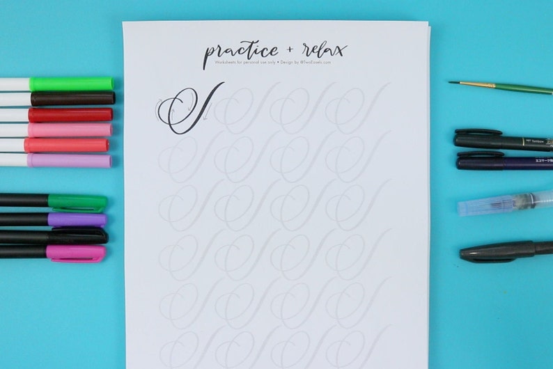 May include: A white sheet of paper with the text "practice + relax" at the top. The paper has a grid of cursive "S" shapes for practicing calligraphy. There are various pens and markers on a blue surface next to the paper.