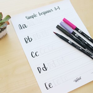 Pu&ograve; includere: Un foglio di carta bianco con carta rigata e il testo "Simple beginner A-Z" in alto. Le lettere "Aa", "Bb", "Cc", "Dd" ed "Ee" sono scritte in corsivo sulla carta. Quattro pennelli neri con tappi rosso, viola e rosa sono appoggiati sulla carta.