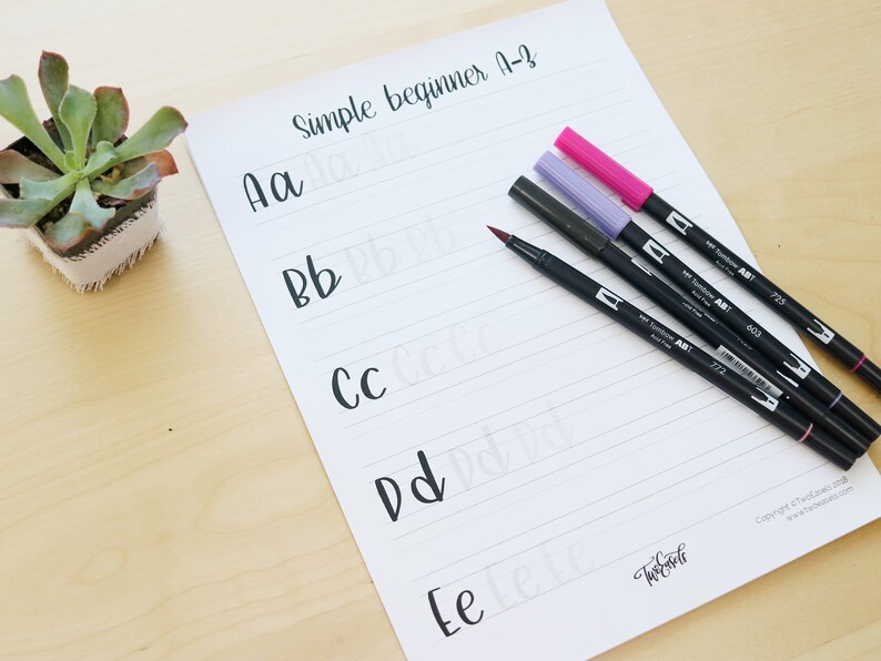 Pu&ograve; includere: Un foglio di carta bianco con carta rigata e il testo "Simple beginner A-Z" in alto. Le lettere "Aa", "Bb", "Cc", "Dd" ed "Ee" sono scritte in corsivo sulla carta. Quattro pennelli neri con tappi rosso, viola e rosa sono appoggiati sulla carta.