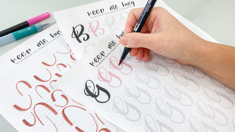 K&ouml;nnte beinhalten: Eine Person &uuml;bt Kalligraphie mit einem schwarzen Pinselstift auf einem Blatt Papier mit grauen und rosa Hilfslinien. Das Papier tr&auml;gt den Text "keep me busy" oben. Die Person &uuml;bt das Schreiben der Buchstaben "B" und "G".
