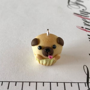 Pu&ograve; includere: Un piccolo ciondolo a forma di cupcake beige a forma di carlino con orecchie marroni, occhi neri e una lingua rosa che sporge. Il ciondolo ha un anello d'argento per appenderlo.
