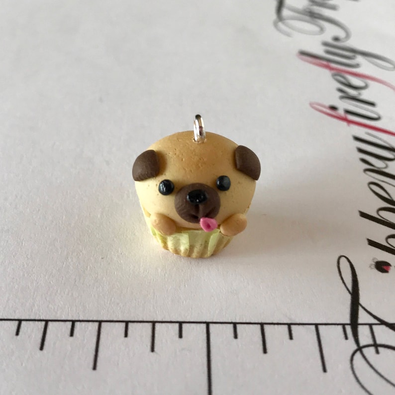 Pu&ograve; includere: Un piccolo ciondolo a forma di cupcake beige a forma di carlino con orecchie marroni, occhi neri e una lingua rosa che sporge. Il ciondolo ha un anello d'argento per appenderlo.