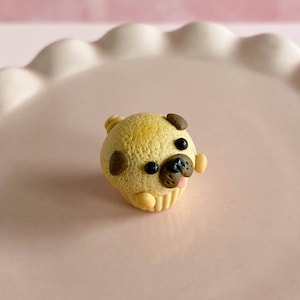 Pu&ograve; includere: Un piccolo cupcake a forma di carlino marrone e beige con occhi neri e una lingua rosa.