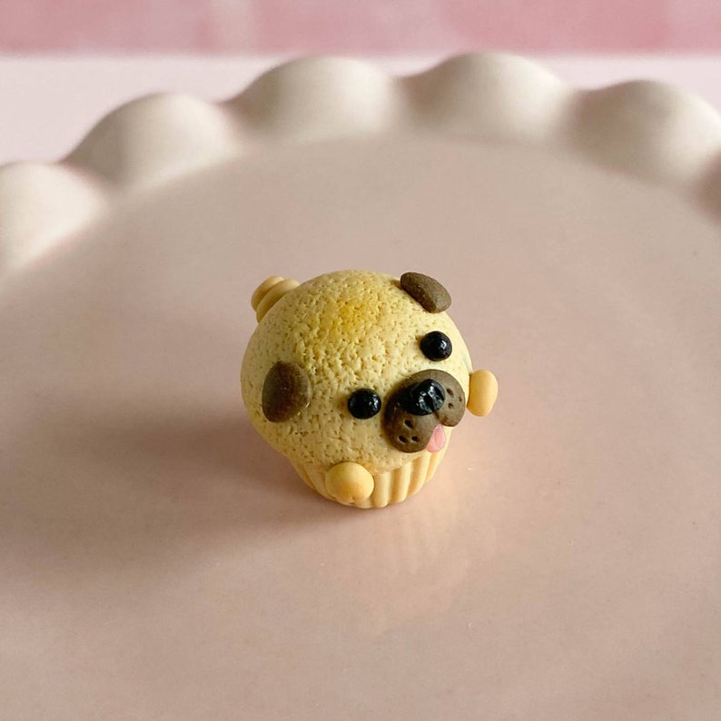 Pu&ograve; includere: Un piccolo cupcake a forma di carlino marrone e beige con occhi neri e una lingua rosa.