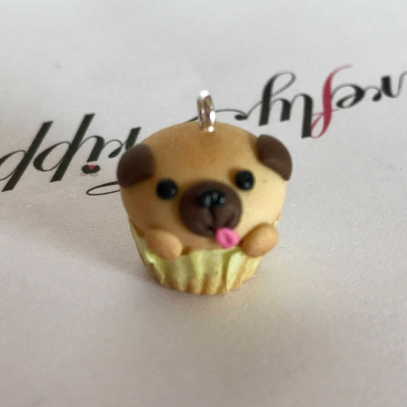 Pu&ograve; includere: Un piccolo ciondolo a forma di cupcake con un carlino marrone e beige, con la lingua rosa che sporge. Il ciondolo &egrave; fatto di pasta polimerica e ha un anello d'argento per appenderlo.