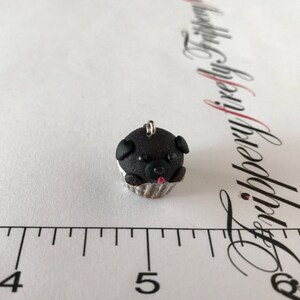 Pu&ograve; includere: Un ciondolo a forma di cupcake nero con un muso di cane, occhi neri e una lingua rosa. Il cupcake &egrave; in un pirottino da cupcake argentato.