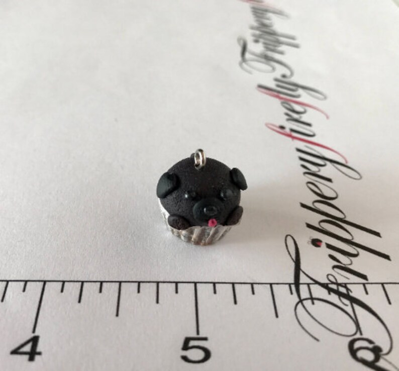 Pu&ograve; includere: Un ciondolo a forma di cupcake nero con un muso di cane, occhi neri e una lingua rosa. Il cupcake &egrave; in un pirottino da cupcake argentato.