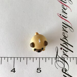 Pu&ograve; includere: Una piccola statuina di carlino beige con orecchie marroni e occhi neri. La statuina &egrave; realizzata in pasta polimerica e ha un anello di filo in cima per appenderla.