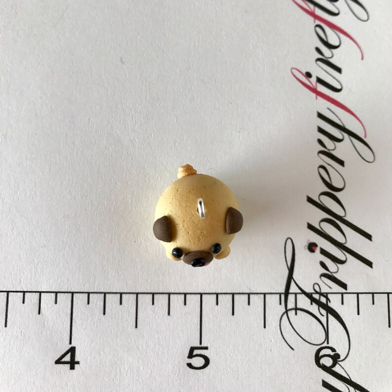 Pu&ograve; includere: Una piccola statuina di carlino beige con orecchie marroni e occhi neri. La statuina &egrave; realizzata in pasta polimerica e ha un anello di filo in cima per appenderla.