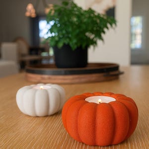 Puede incluir: Dos candelabros con forma de calabaza, uno blanco y otro naranja, sobre una mesa de madera. Cada soporte tiene una vela encendida en su interior. Una planta en una maceta negra y una bandeja de madera en el fondo.