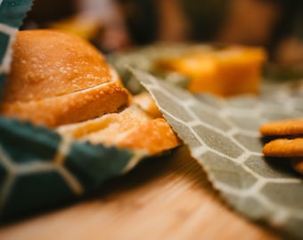 May include: A close-up of a loaf of bread wrapped in a green and white patterned reusable food wrap. The bread is sliced and the edges are visible. There are also some crackers on a similar patterned wrap in the background.