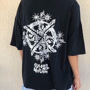 以下が含まれることがあります： 背面に白いグラフィックデザインが施された黒いTシャツ。デザインは、尖った要素と「Ocean in Motion」のテキストを含む円形のパターンを特徴としています。Tシャツは半袖でゆったりとしたフィット感です。
