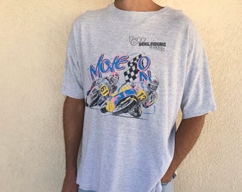 May include: A light gray t-shirt with a graphic of motorcycle racers and the words "Move On". The shirt is paired with light blue denim shorts. The overall style is casual and vintage.