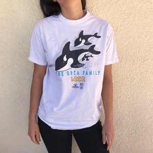 May include: White t-shirt with a graphic of an orca family and the text "THE ORCA FAMILY LORO PARQUE". The orcas are black and white, with blue and yellow text. The shirt has short sleeves.