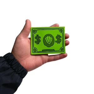 May include: A green patch resembling a dollar bill, featuring dollar signs and a central seashell design. The patch has a rectangular shape with a green border and is held in a hand against a white background.