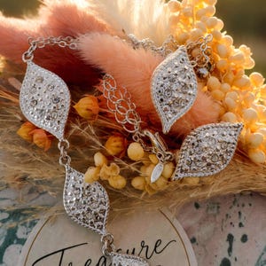 Peut inclure: Un collier argenté composé de trois pendentifs en forme de feuille incrustés de cristaux transparents. Le bijou est présenté sur une carte circulaire portant l'inscription Treasure Lane Costume Jewellery et le slogan little details, big impact.
