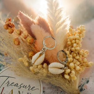 May include: Two silver-toned hoop earrings featuring white cowrie shell charms. The earrings are displayed against a small bouquet of dried beige and pink grasses and flowers, next to a circular tag labelled Treasure LANE COSTUME JEWELLERY.