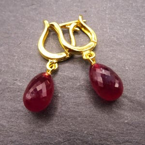 May include: A pair of gold-colored hoop earrings with teardrop-shaped, faceted, deep red gemstones. The earrings are displayed on a dark gray surface, showcasing the jewelry's design and color.
