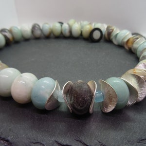 Può includere: Una collana realizzata con perline di colore blu chiaro e marrone con accenti argentati. Le perline sono rotonde e hanno una texture liscia. La collana è infilata su una catena d'argento.