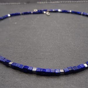 Può includere: Una collana girocollo composta da piccole perline quadrate blu scuro con accenti color argento. Le perline sono infilate insieme per formare una collana. La collana è presentata su una superficie grigio scuro.