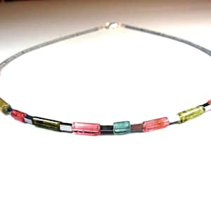 May include: A delicate necklace featuring a silver chain with a series of colorful, rectangular beads. The beads are in shades of pink, green, and blue, interspersed with small, metallic spacers. The necklace is designed to be worn around the neck.